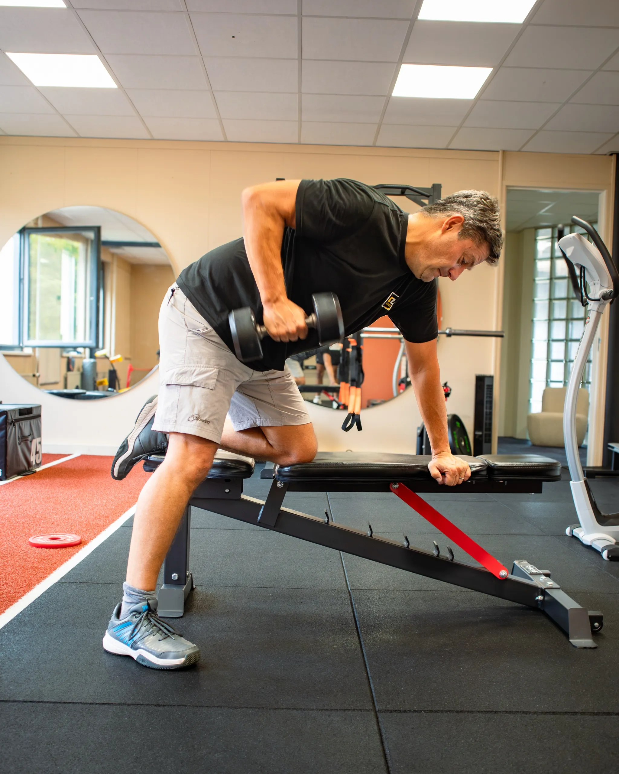 Een cliënt roeit met een dumbbell op een bankje, met de ronde spiegel van de studio achter hem