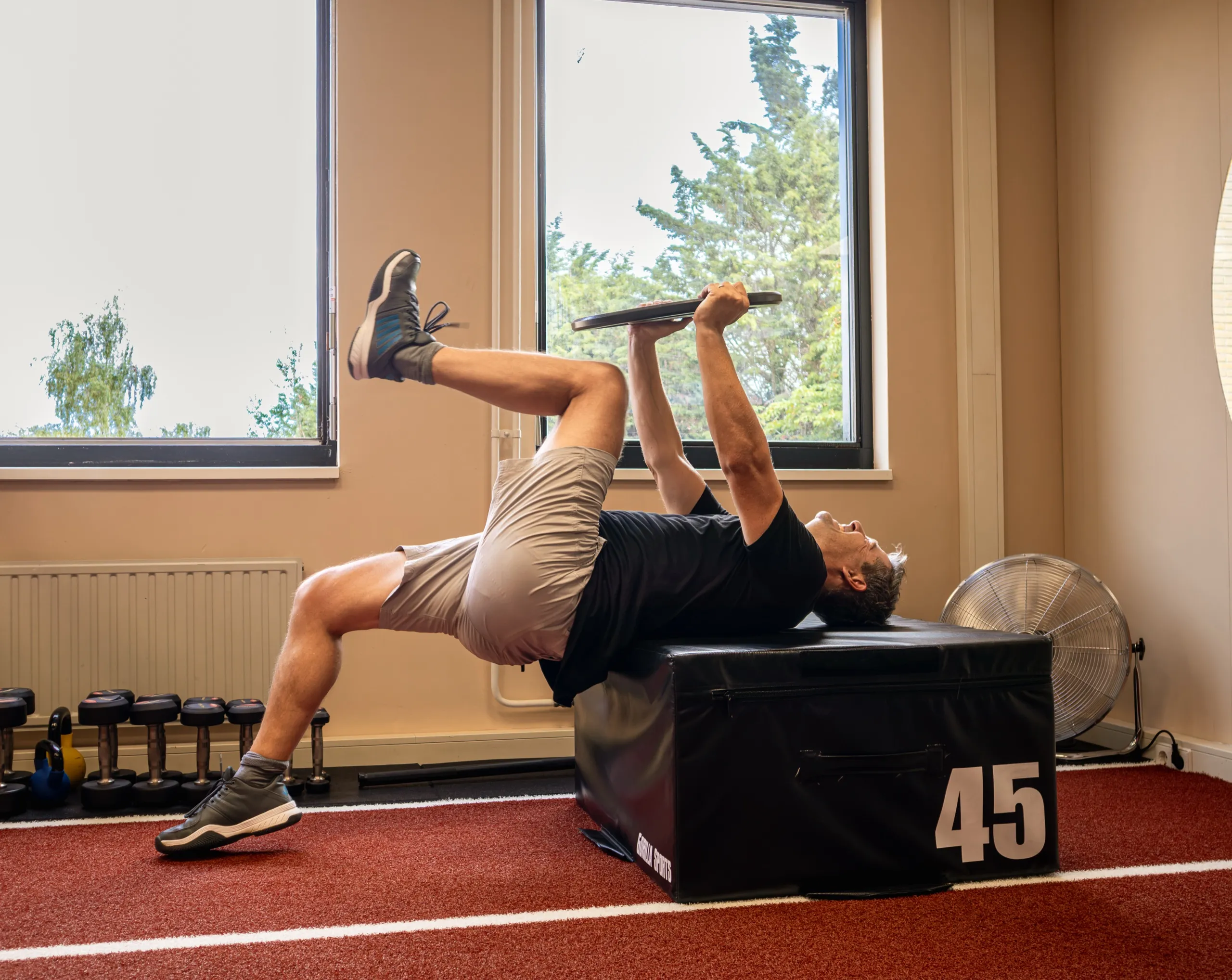Een cliënt doet een hip thrust op een plyobox voor de ramen van de studio, met bomen buiten