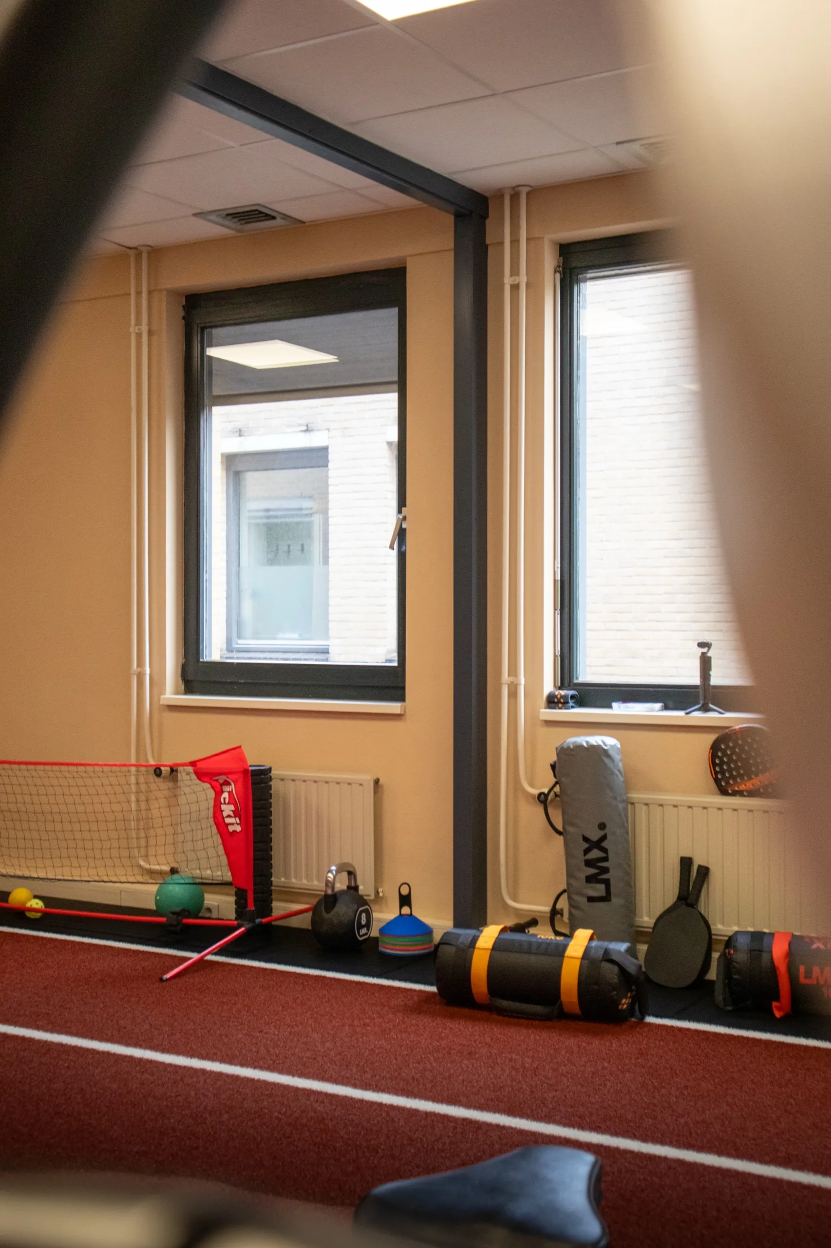Hoek van de studio van Bekafit bij de ramen, met een voetbalnetje, een kettlebell en powerbags op de rode sprintbaan
